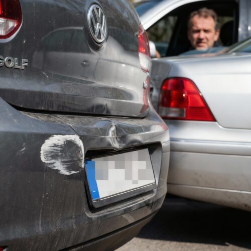Nahaufnahme der hinteren Stoßstange eines stehenden Autos, die neuen Aufprallschaden links direkt neben einer alten Delle zeigt, während das verursachende Auto noch am Kontaktpunkt steht.