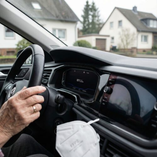 Das digitale Armaturenbrett eines VW Polo zeigt eine extrem geringe Kilometerzahl; im Bild sind die Hand der älteren Fahrerin und ein Mundschutz.